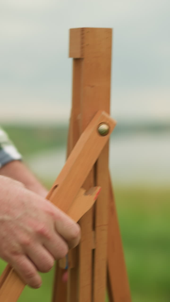 A close-up shot of a man in a plaid shirt holding and setting up a wooden tripod outdoors. In the background, a woman in a white dress is blurred, creating a focus on the man s actions and the tripod