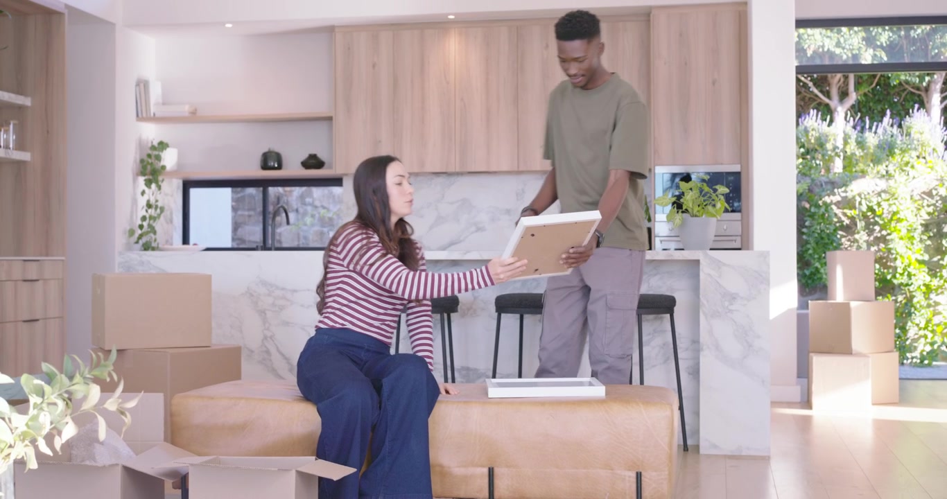 Diverse couple unpacking framed pictures and decorating after moving into open-plan living area