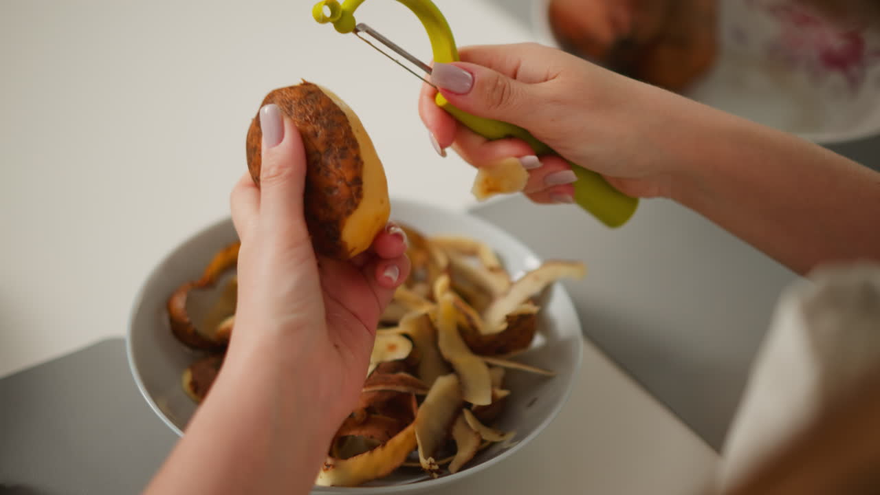Hand view of person peeling potato using peeler with other hand holding potato firmly, over plate filled with discarded skin and blurred unpeeled potatoes in background on kitchen counter