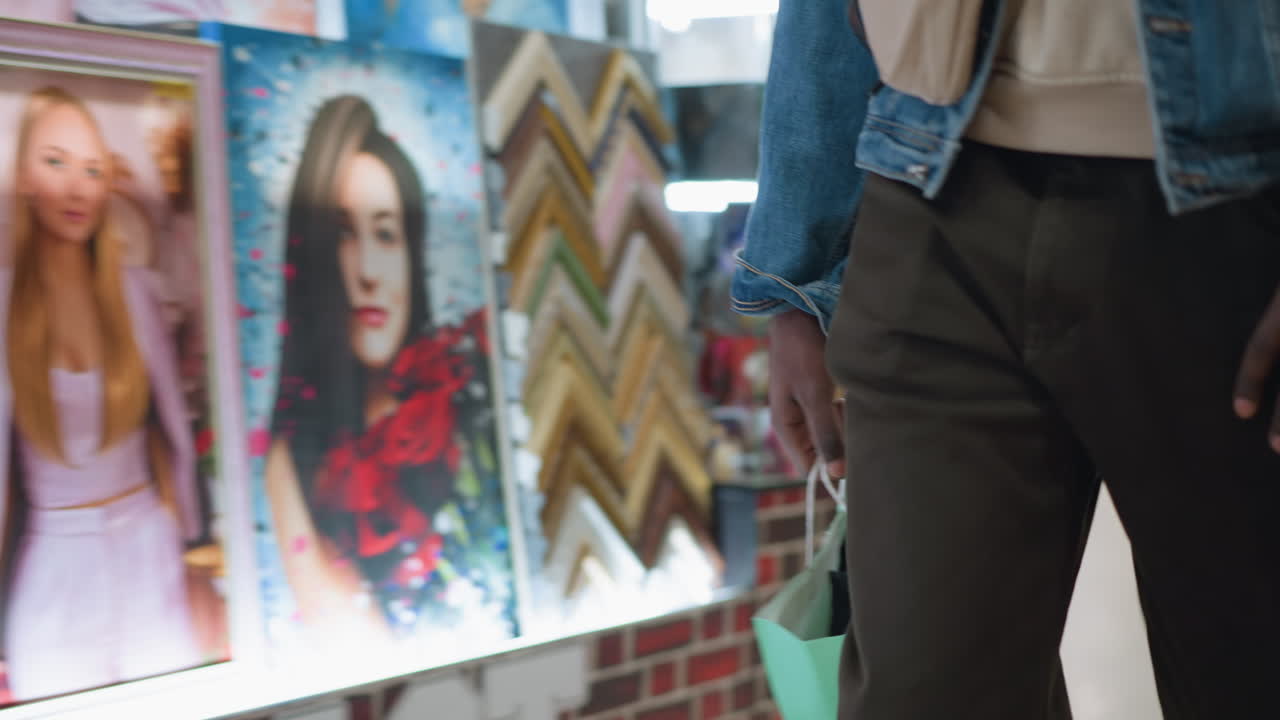 close shot of male shopping in mall carrying green paper bag by denim jacket and pants hand gripping handle against blurred retail interior with bright lights and bokeh creating candid consumer mood