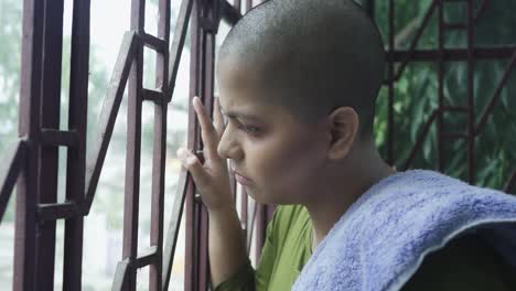 una joven india enferma y calva con cáncer mirando afuera a través de la ventana y molesta