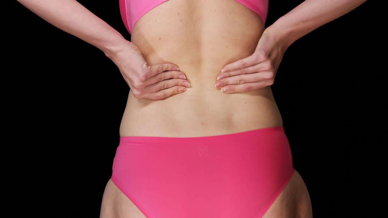 mujer joven con tanga rosa y sostén tiene dolor de espalda y se masajea
