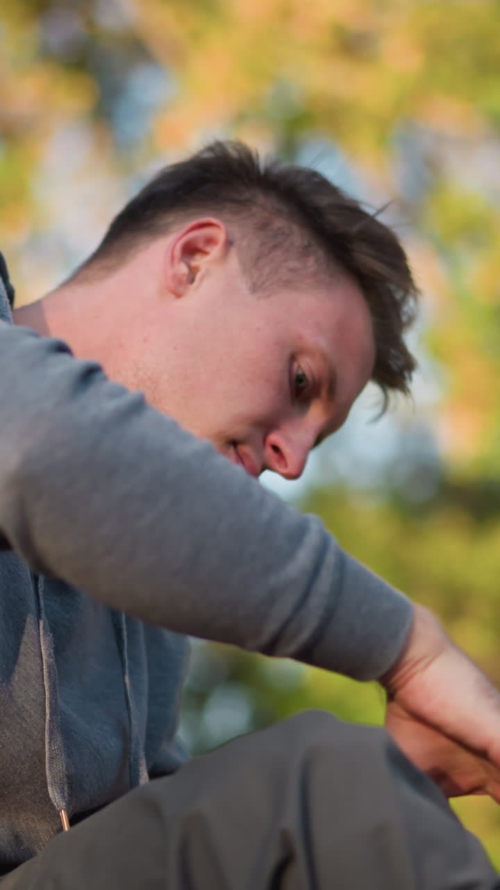 회색 후디를 입은 젊은이의 가까운 은 그가 검은색과 오렌지색 스트을 들고있는 동안 집중적으로 초점을 맞추고, 흐릿한 자연 야외 배경에 대해 깊은 생각이나 집중으로 나타납니다.