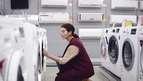 una hermosa chica morena baja para abrir la puerta de una lavadora en una tienda de electrodomésticos, buscando el equipo doméstico perfecto.