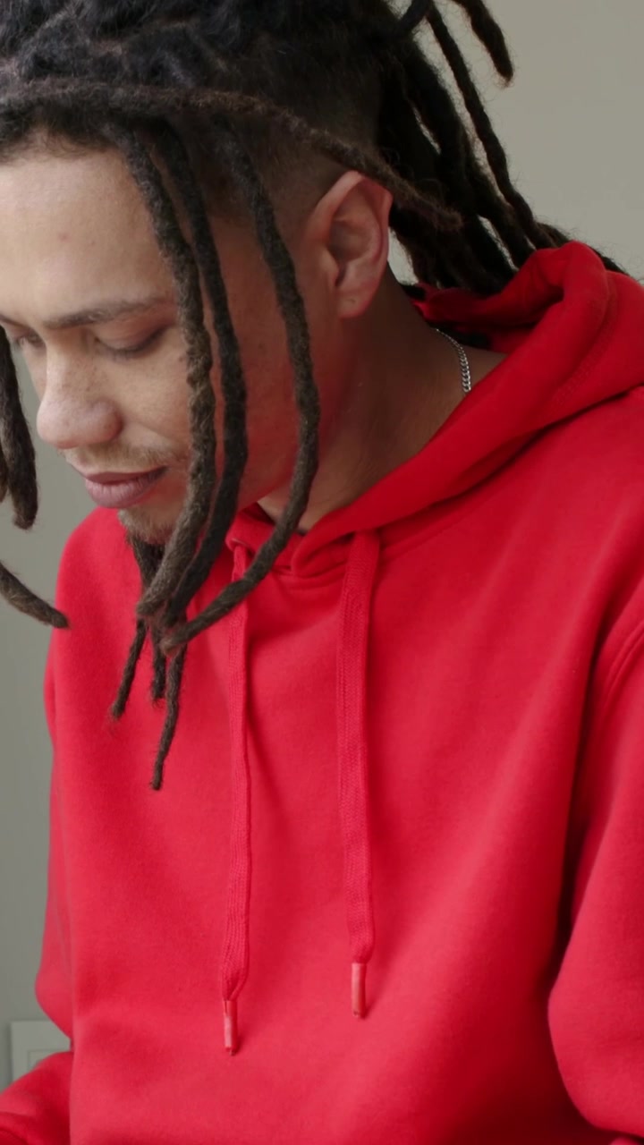 Video verticale: Un uomo con i dreadlock, con una felpa rossa con cappuccio, che controlla sotto l'inquadratura in una stanza grigia.