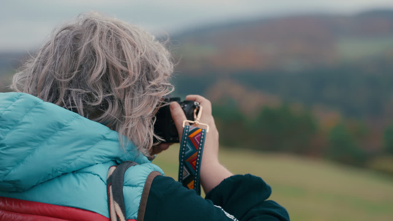 close up de mujer de cabello gris fotógrafa desde atrás tomando fotos de coloridas naturalezas de otoño naranja con su cámara y mirando a su alrededor durante un día frío y ventoso en cámara lenta