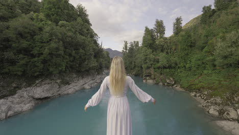 modelo rubia con vestido blanco levantando los brazos lentamente mientras mira el río azul vibrante