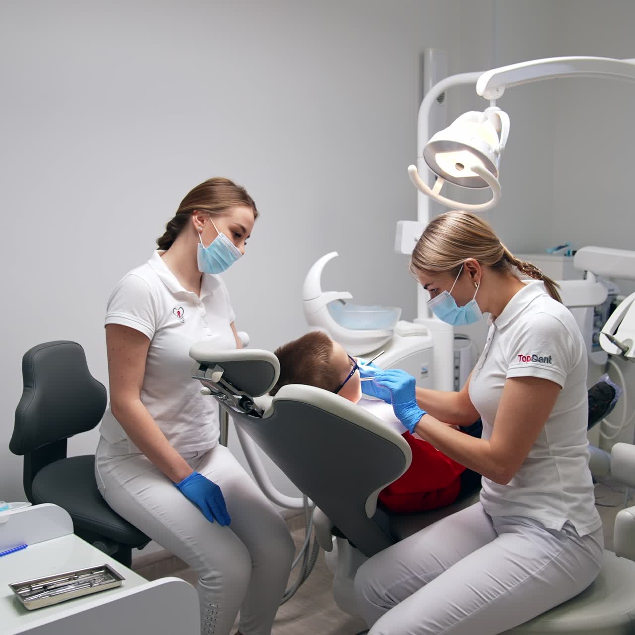 Lady dentist in white uniform applying metal instruments in her work. Professional is examining the little boy sitting in dental chair