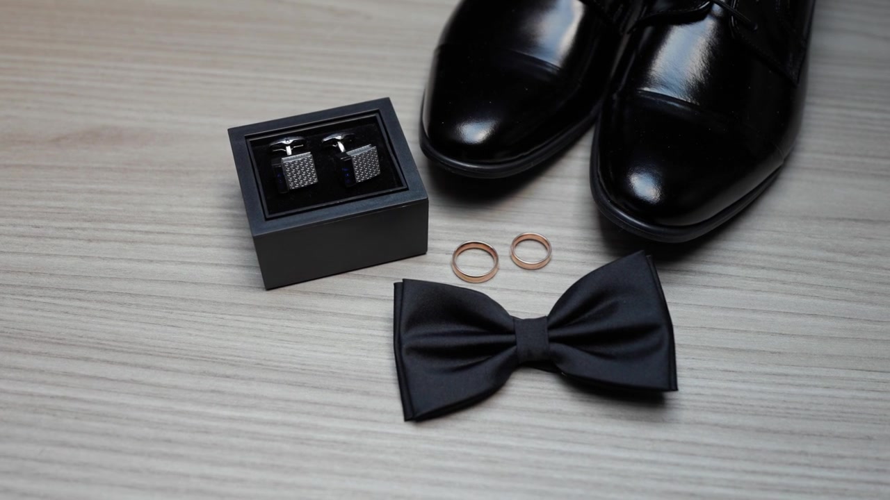 Stylish close-up shot of groom’s black shoes, wedding rings, bow tie, and cufflinks on wood surface. Soft lighting highlights luxury and timeless elegance for wedding themes.