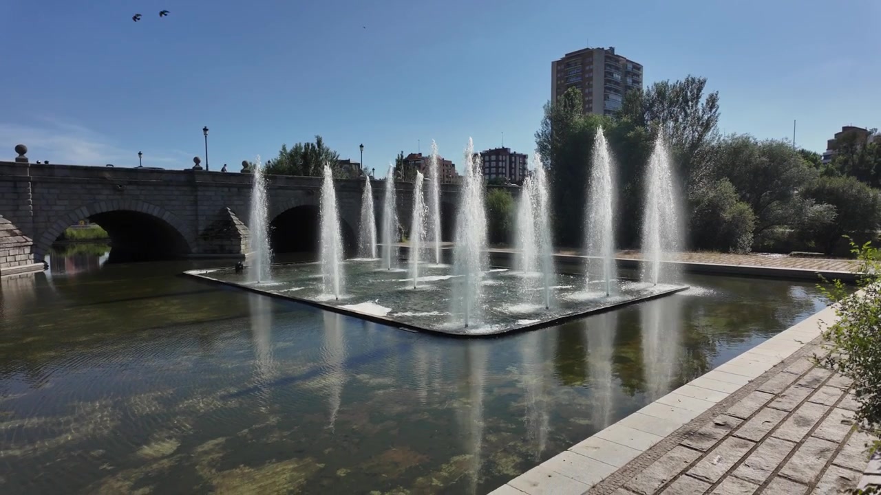 Elegantes fuentes de agua se elevan en cámara lenta junto al histórico puente de Segovia sobre el río Manzanares en Madrid, España, con edificios modernos y un cielo azul claro de fondo.