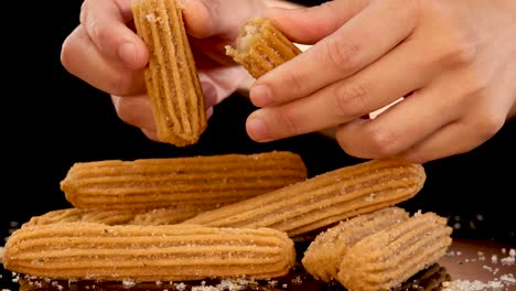 close-up of hands breaking a sugar-dusted churro stick over a stack of churros on a dark background, with warm studio lighting and steady camera