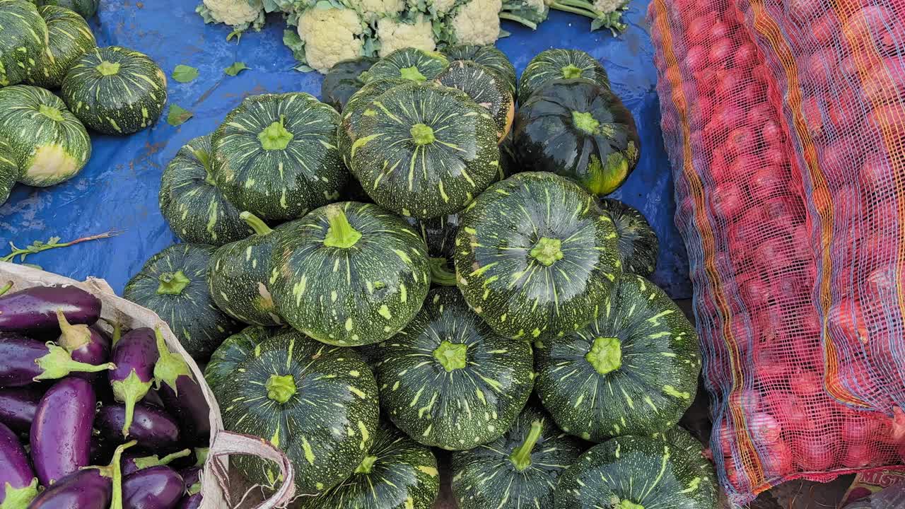 Camera circles around a neatly stacked pile of green pumpkins with yellow speckles, surrounded by other market vegetables on a blue sheet in a vibrant outdoor setup
