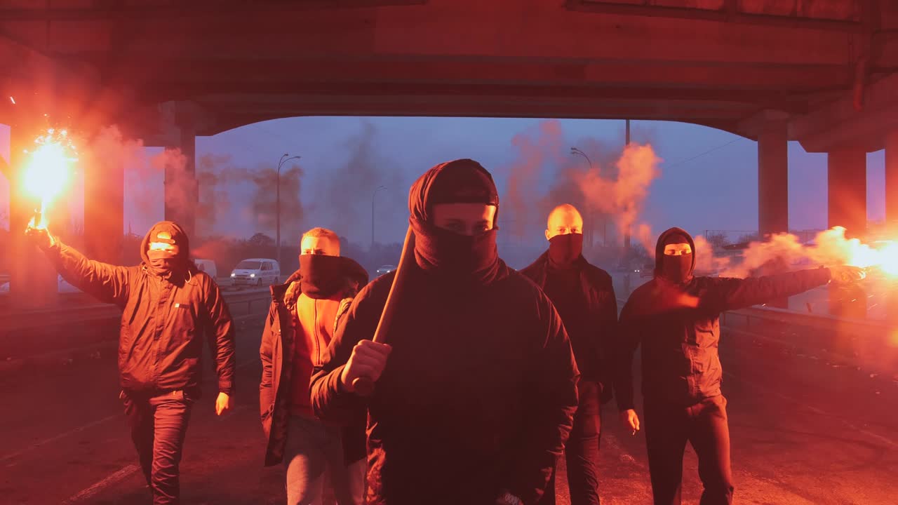 Group of young men in balaclavas with red burning signal flare walking on the road under the bridge, slow motion