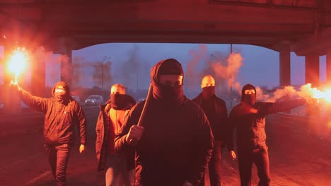 group of young men in balaclavas with red burning signal flare walking on the road under the bridge, slow motion