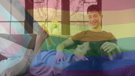 animation of rainbow flag over happy diverse gay male couple relaxing on couch using smartphone