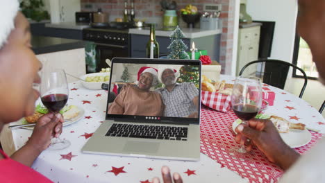 african american couple with wine using laptop for christmas video call with couple on screen