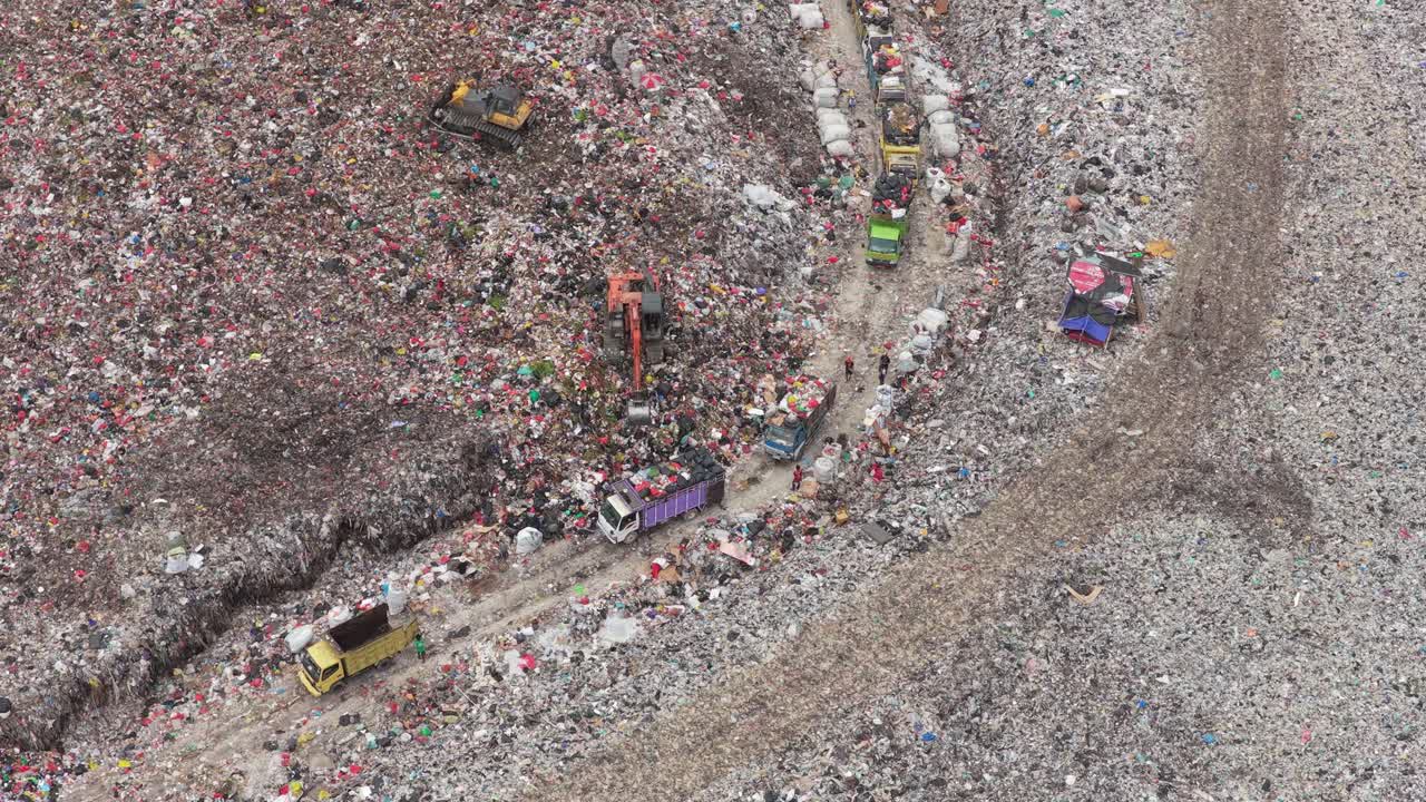 Vista aerea dall'alto di macchinari pesanti e code di camion della spazzatura che scaricano plastica e rifiuti domestici in una vasta discarica, illustrando l'inquinamento, l'iperconsumo e la crisi della gestione dei rifiuti.