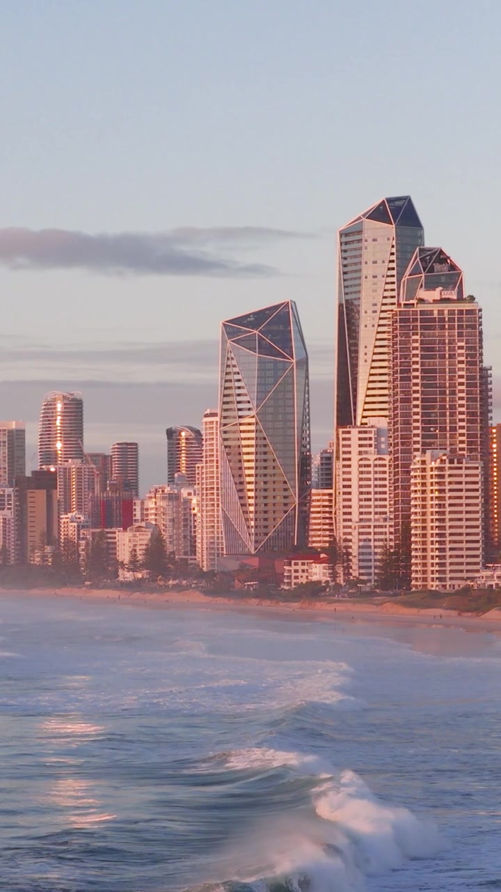 Vertical drone shot of modern city skyscrapers along a beach with rolling ocean waves.