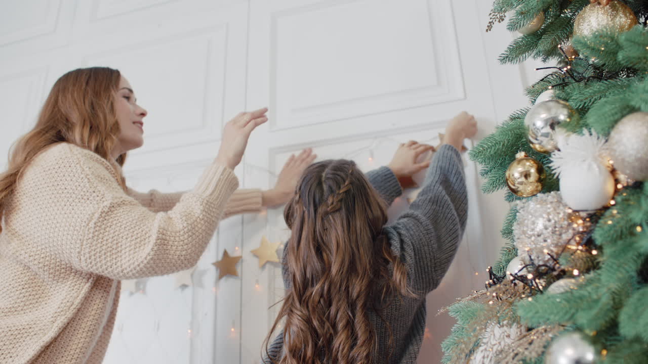 Smiling mom and teen daughter putting garland on wall in living room.
