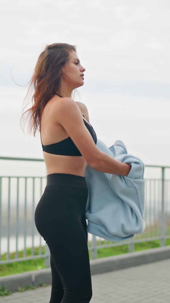 Lady standing in front of iron fence, removing her blue hoodie under sunny weather with a river background. Ideal for fitness, outdoor, and lifestyle moments in a bright, active environment