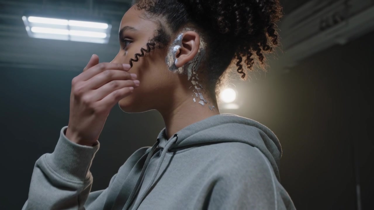 Levá ruka se zvedá, žena v šedé mikině odhaluje stříbrnou náušnici, drží pramen vlasů s protisvětlem ve studiu.