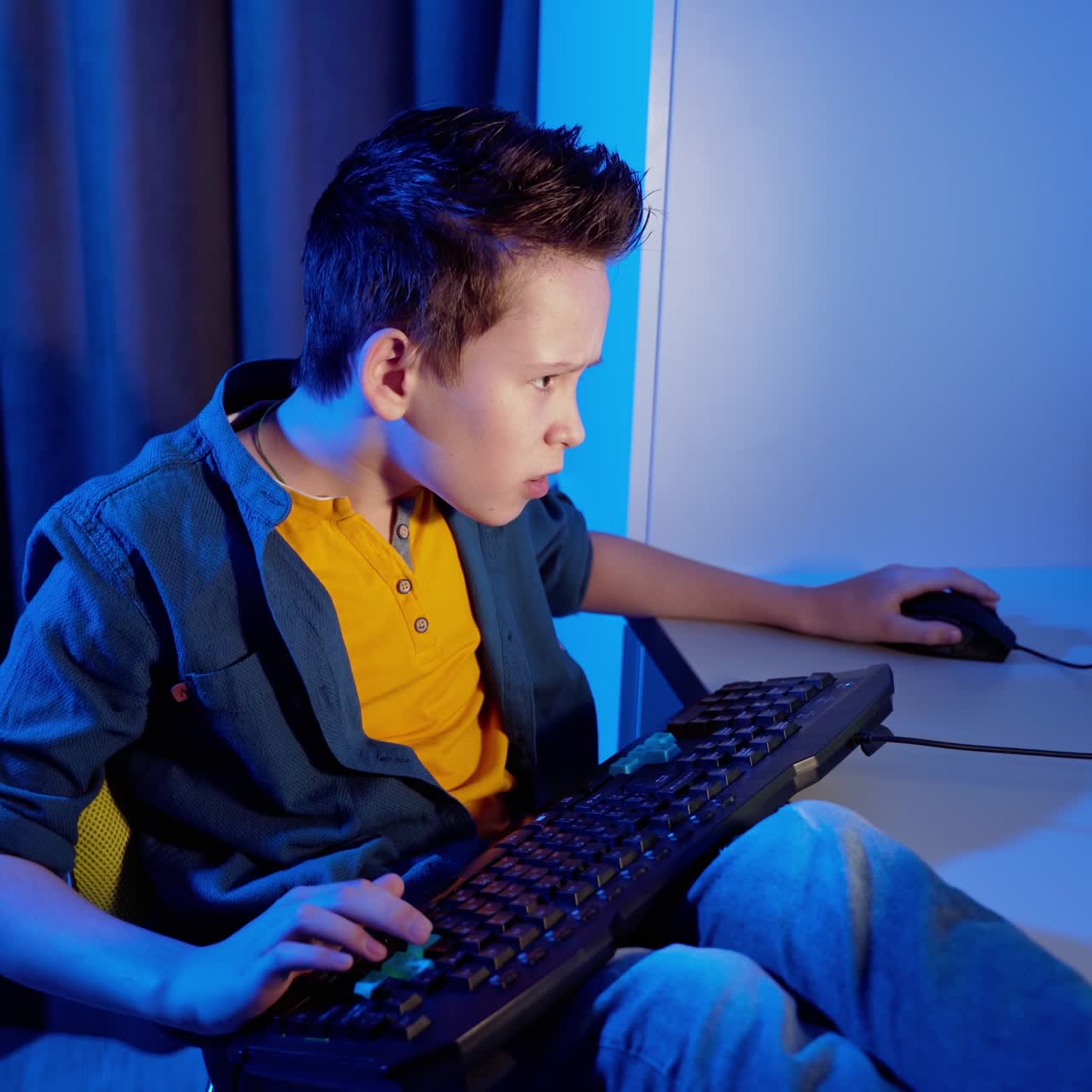 Children's entertainment. Boy is playing video games at home. Teenage boy sitting at the desk with a keyboard on legs and concentrated on online games on his computer.