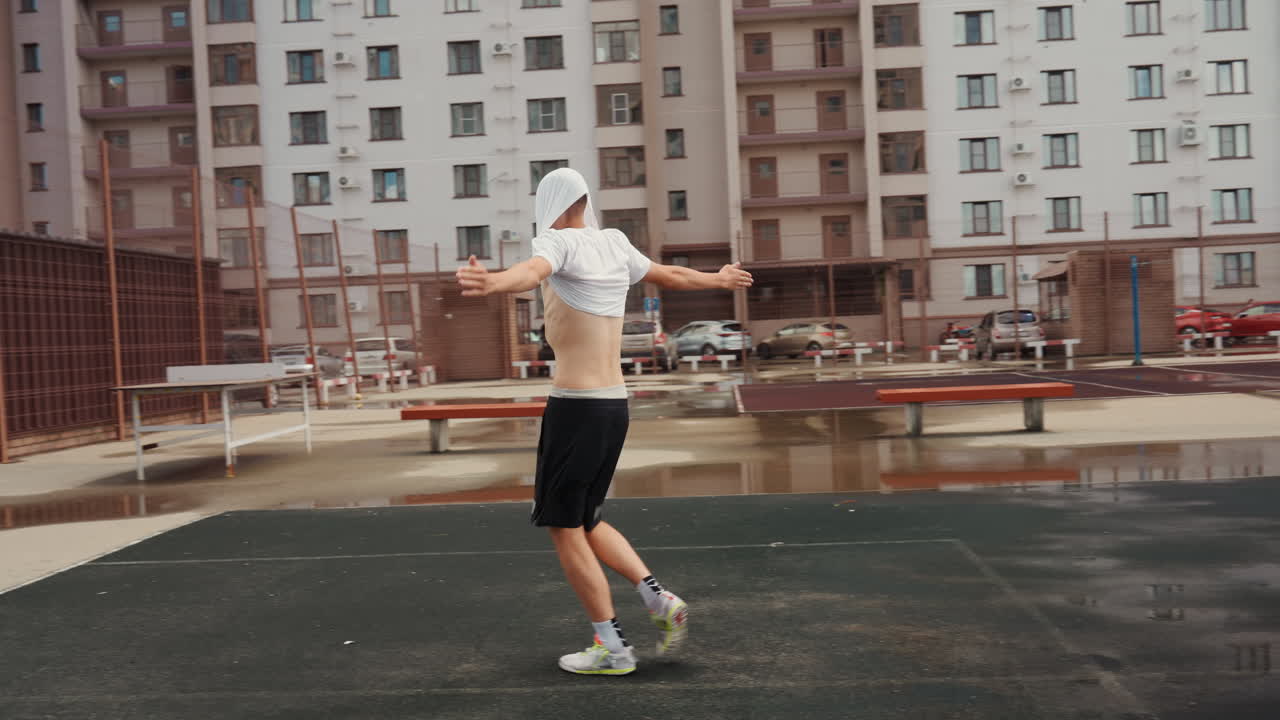 young man warming up after rain in empty construction courtyard with soccer ball, puddles, apartment blocks, overcast sky. removes shirt and practices dribble and freestyle moves on wet asphalt