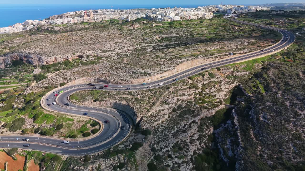 Aerial view of a winding serpentine road cutting through Mellieha's rocky terrain, with the city, Mediterranean Sea, and coastline creating a stunning backdrop under a clear blue sky