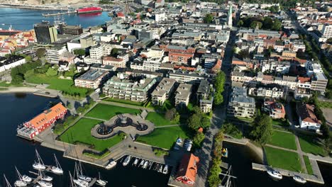 aerial view of kristiansand in norway featuring a bustling waterfront, modern buildings, and lush green parks on a sunny day. boats and ships fill the harbor, enhancing the scenic beauty