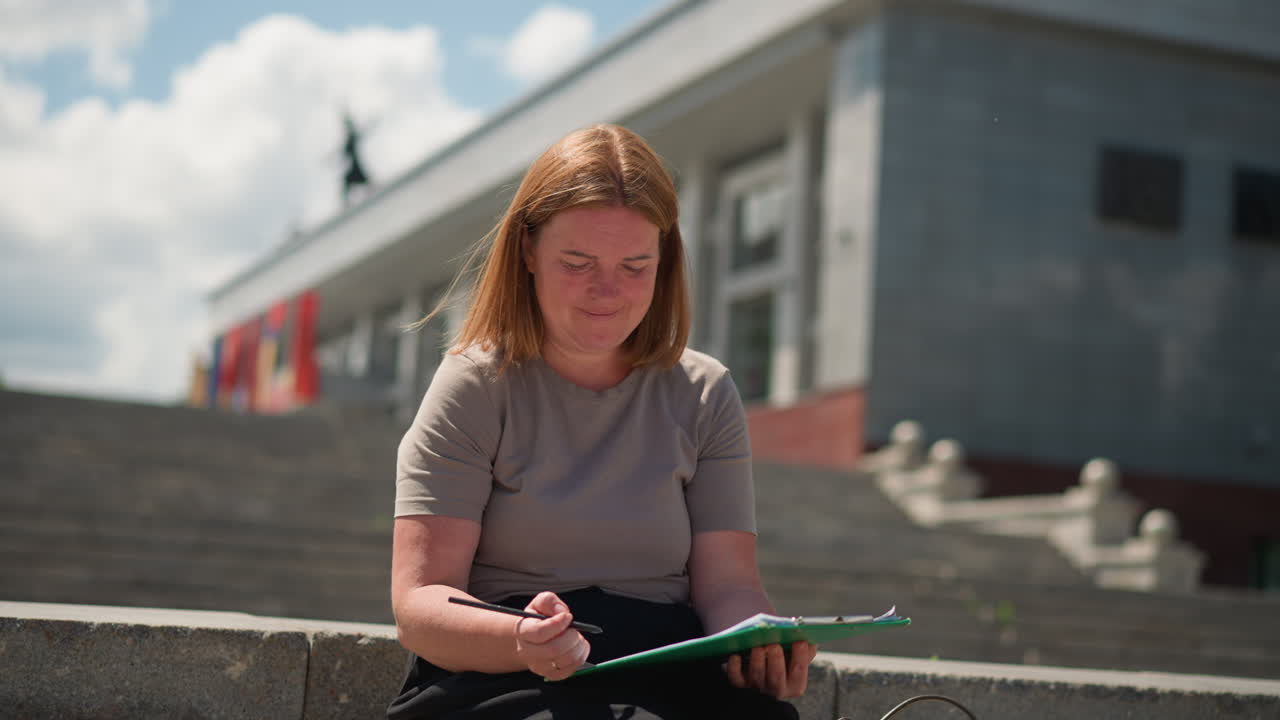 Student sitting alone on stone steps writing on clipboard under warm sunlight, wearing beige top, focused expression, green clipboard in hand, modern building under construction visible in background