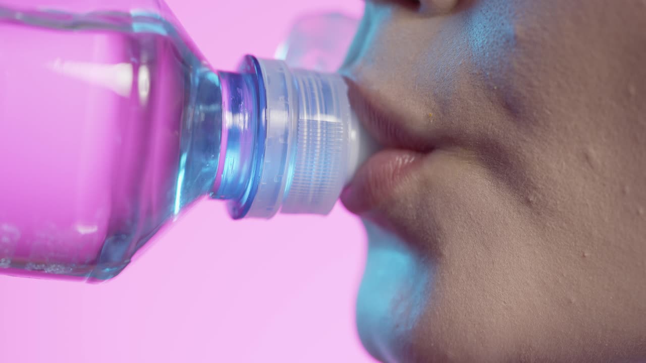 mujer tomando un sorbo de agua de la botella de agua en primer plano, esmalte de uñas femenino y labios de lápiz labial rojo en fondo rosa