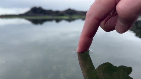 Close-up-of-a-finger-touching-the-reflective-surface-of-a-lake-in-the-mountains