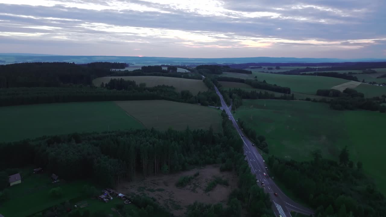 Vej i landskab med forbipasserende biler ved sen solnedgang.