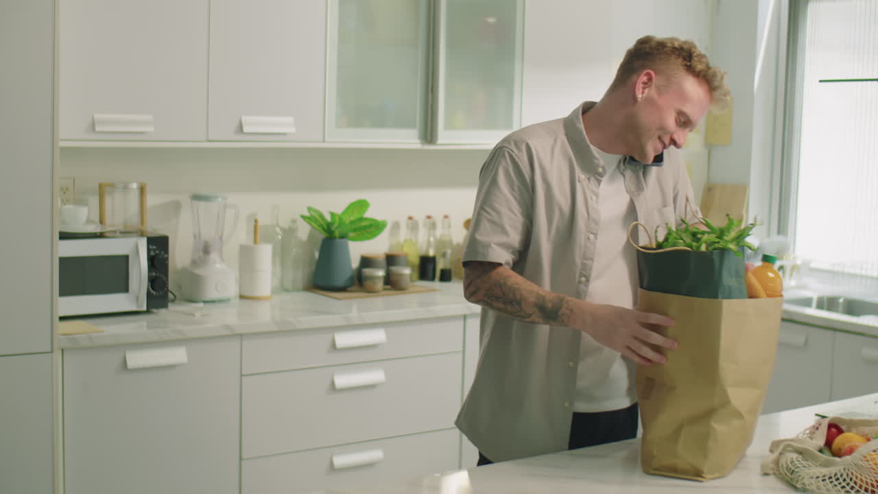 Man Taking Groceries from Bag and Talking on Phone in Kitchen