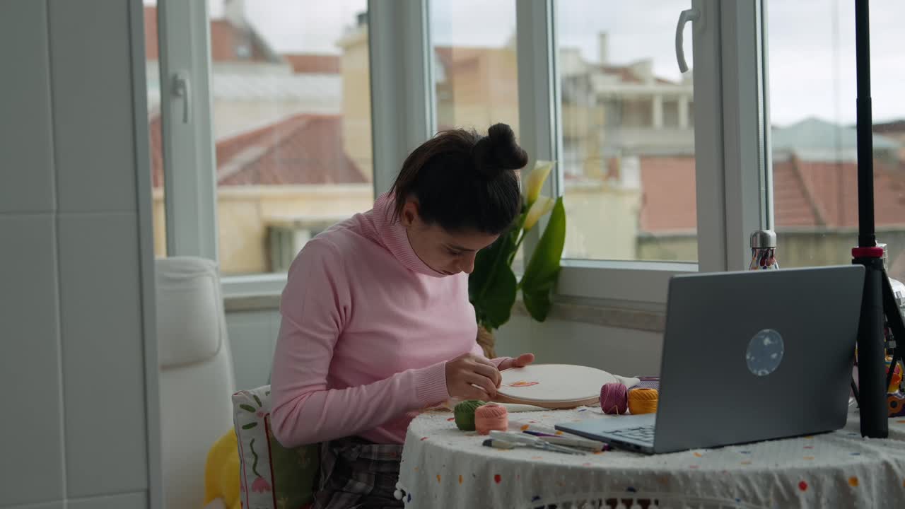 Mujer joven bordando en casa con portátil cerca