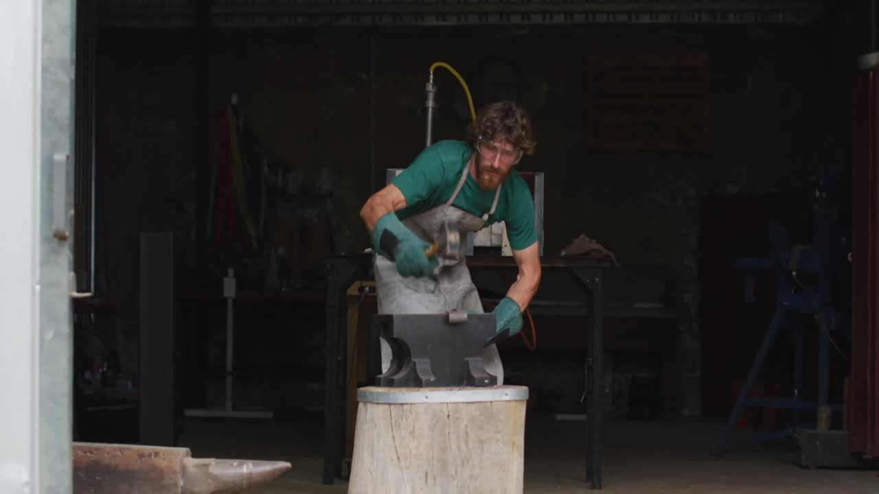 Caucasian male blacksmith wearing safety glasses, hammering hot metal tool on anvil in workshop