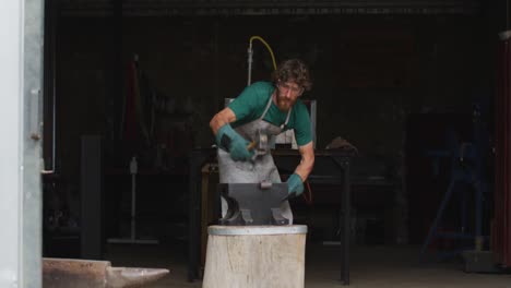 caucasian male blacksmith wearing safety glasses, hammering hot metal tool on anvil in workshop