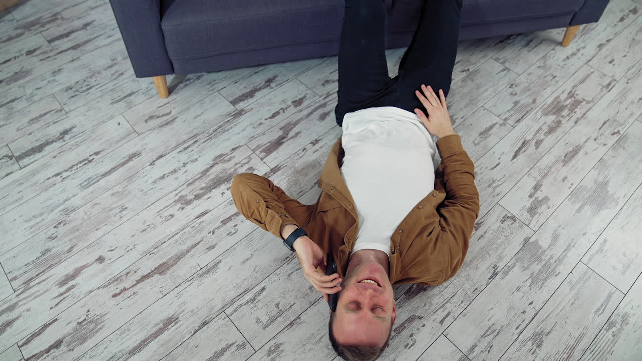Young man talking the phone at home. Happy man having a conversation through the phone while lying on floor near the sofa. Top view. Quarantine.