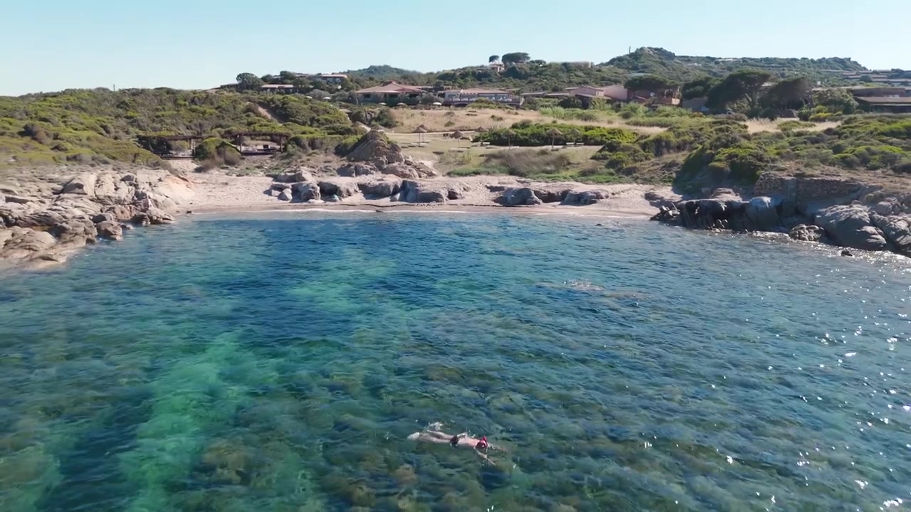 Vista aerea del mare azzurro e limpido, spiaggia rocciosa e ville sulla costa della Sardegna.
