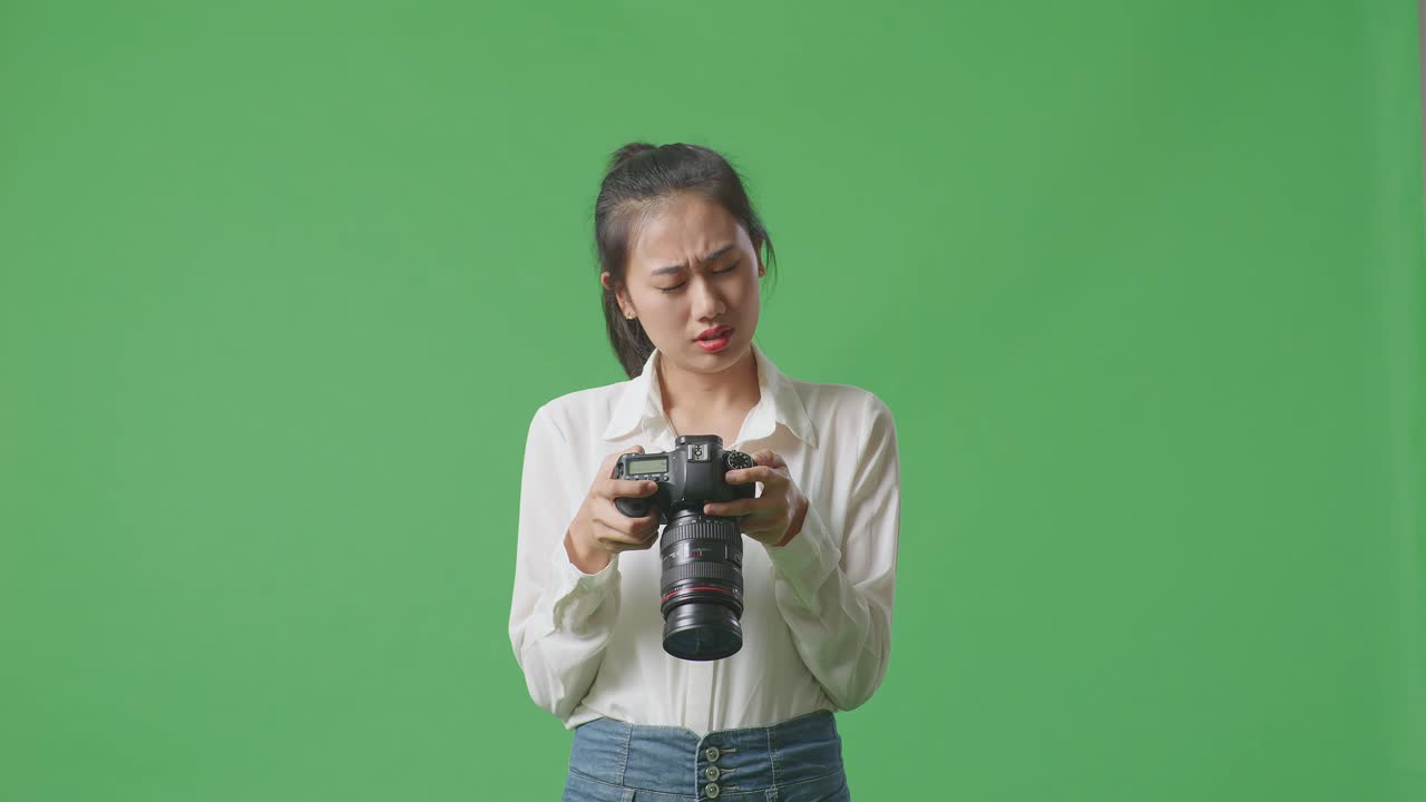 아시아 사진작가 는 스튜디오 에서 녹색 스크린 배경 에 서 있는 동안 카메라 에 있는 사진 을 보고 두통 을 고 있다