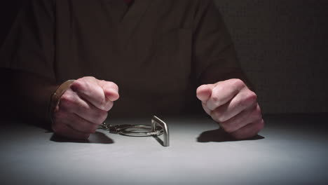 toma de paralaje de primer plano de las manos de un prisionero mientras se sienta esposado a una mesa en una sala de interrogatorios, con las manos apretadas en puños