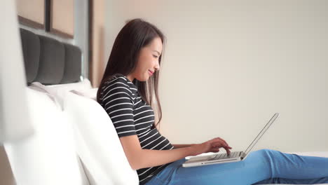 asian woman sitting in bed typing on laptop computer