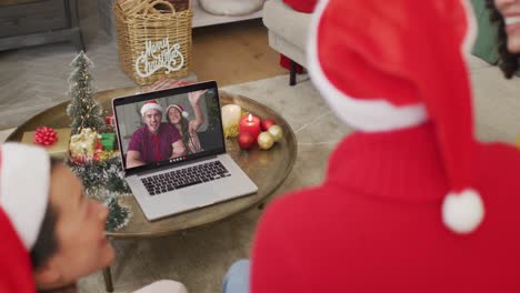 Diverse-family-with-santa-hats-using-laptop-for-christmas-video-call-with-happy-couple-on-screen