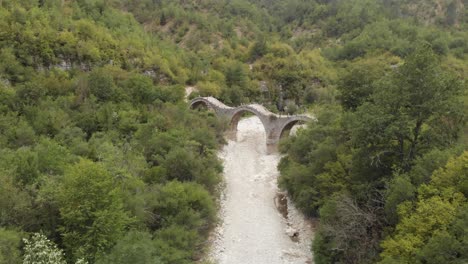 Video-De-Drones-Del-Viejo-Puente-De-Tres-Arcos-De-Piedra-De-La-Región-De-Plakidas-Zagori-Grecia-En-El-Bosque,-Río-Seco-Debajo-Y-Formación-Rocosa-En-La-Parte-Posterior-Panorámica-Izquierda