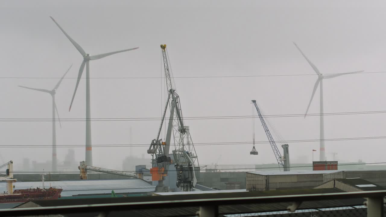 Wide shot of an industrial harbor featuring towering cranes with suspended grab buckets, wind turbines and dense fog covering the skyline