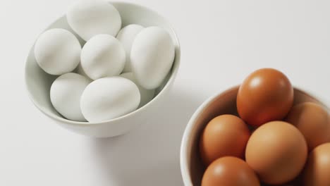 Close-up-of-two-bowls-full-of-brown-and-white-eggs-with-copy-space-on-white-surface