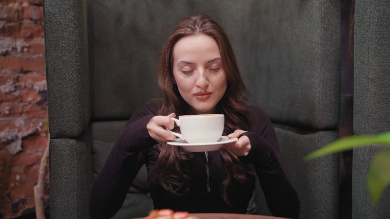 Young woman in dark outfit closes eyes while lifting latte to face, savoring aroma with calm expression. Wooden table holds waffles, white cup, with textured gray booth and brick wall visible