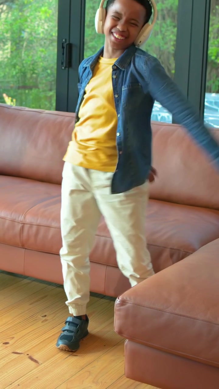 Vertical video: Dancing boy in yellow tee with cream headphones moving to music at home by sofa