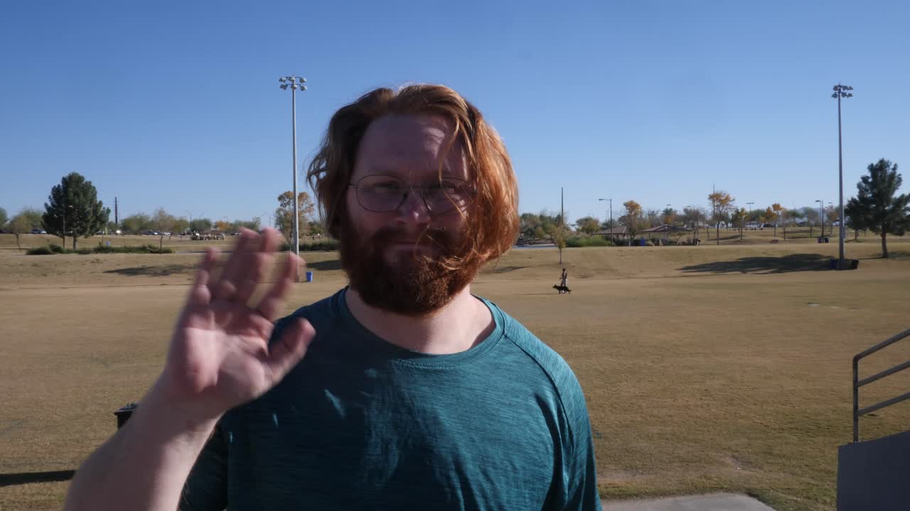 A red haired man waving at the camera in a public park.