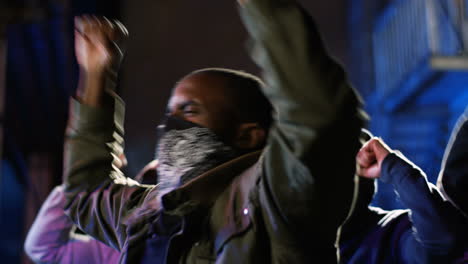 Side-view-of-african-american-man-with-scarf-on-his-face-yelling-with-arms-up-in-a-protest-with-multiethnic-group-of-people-in-the-street